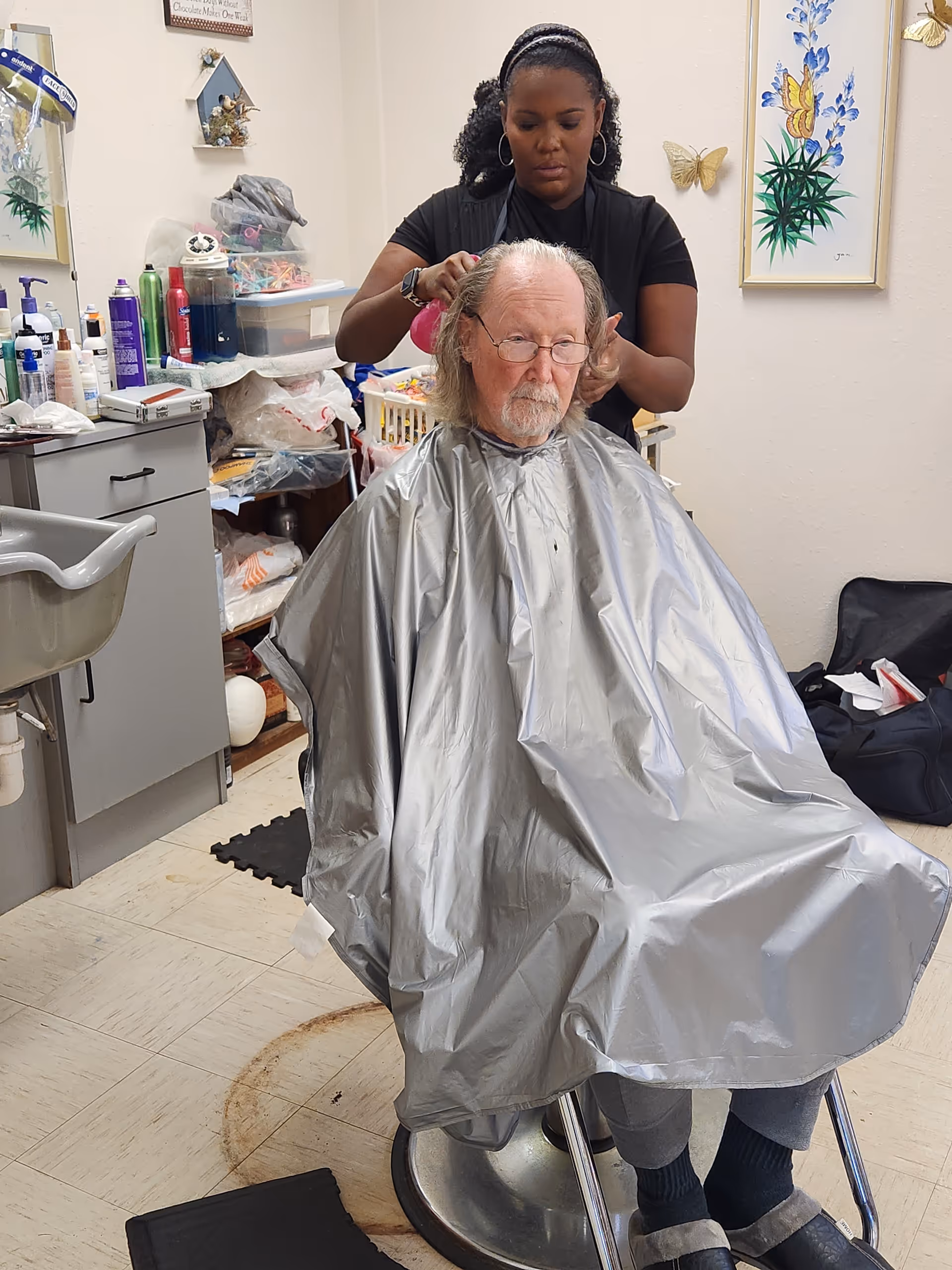 An elderly man with glasses is seated in a salon chair wearing a silver haircut cape while a woman stands behind him styling his hair. The room contains various hair care products and supplies on shelves and counters, with framed artwork and butterfly decorations on the wall.