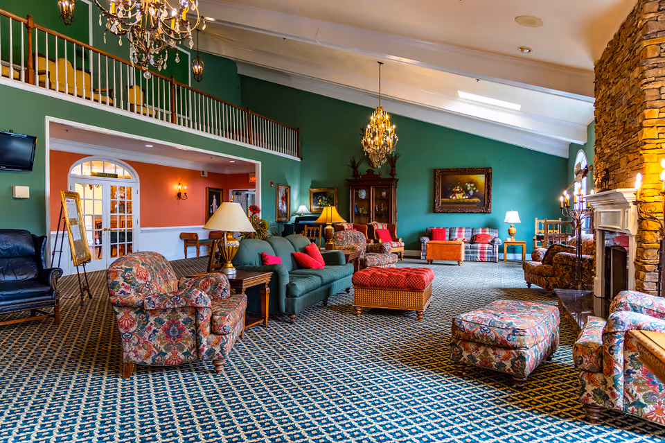 A spacious senior living facility common area with high vaulted ceilings, green walls, and patterned carpet. The room is furnished with various floral and solid-colored sofas, armchairs, and ottomans arranged around a stone fireplace on the right. There are multiple table lamps and chandeliers providing warm lighting, and framed artwork adorns the walls. A balcony railing is visible above an open doorway leading to another room with peach-colored walls.