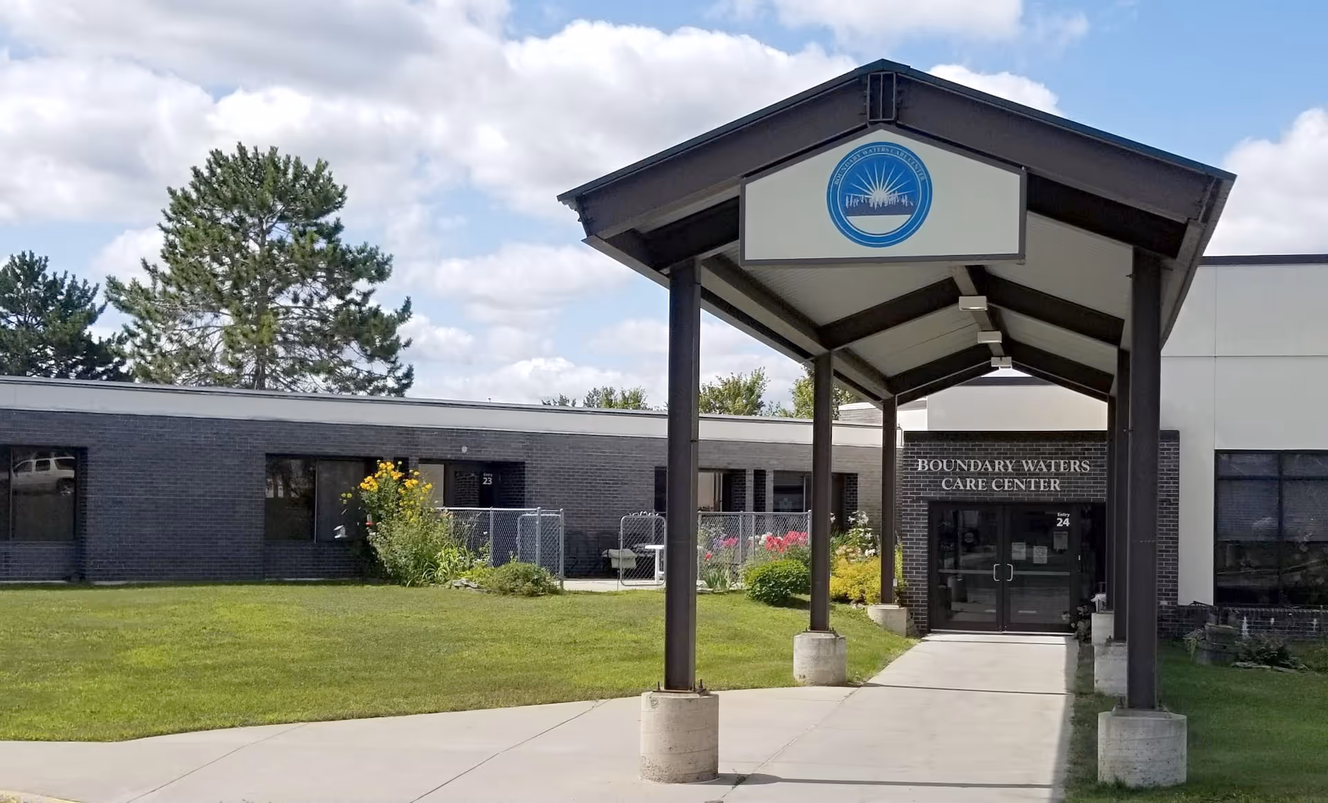 Exterior view of Boundary Waters Care Center showing a covered entrance with a walkway leading to glass double doors. The building is single-story with dark brick and light-colored walls. There is a grassy lawn with some bushes and flowers, and a large tree in the background under a partly cloudy sky.