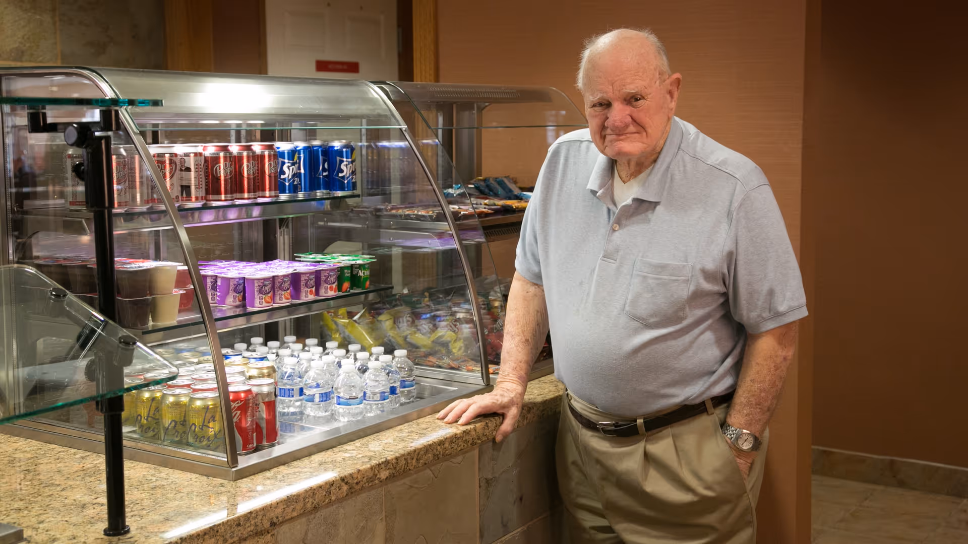 An elderly man wearing a light gray polo shirt and beige pants stands next to a snack and beverage display case in an indoor setting. The display case contains various canned sodas, bottled water, yogurt cups, and snack bags. The background features a brown wall and tiled floor.