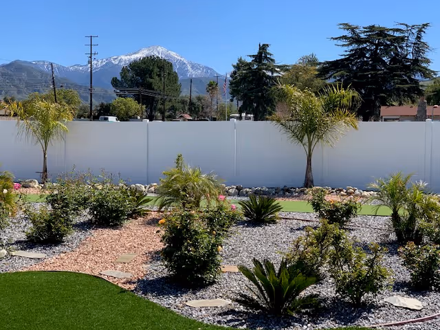 A landscaped outdoor garden area with various small palm trees, bushes, and plants arranged on gravel and mulch beds. A white fence runs along the background with tall trees and a snow-capped mountain visible in the distance under a clear blue sky.