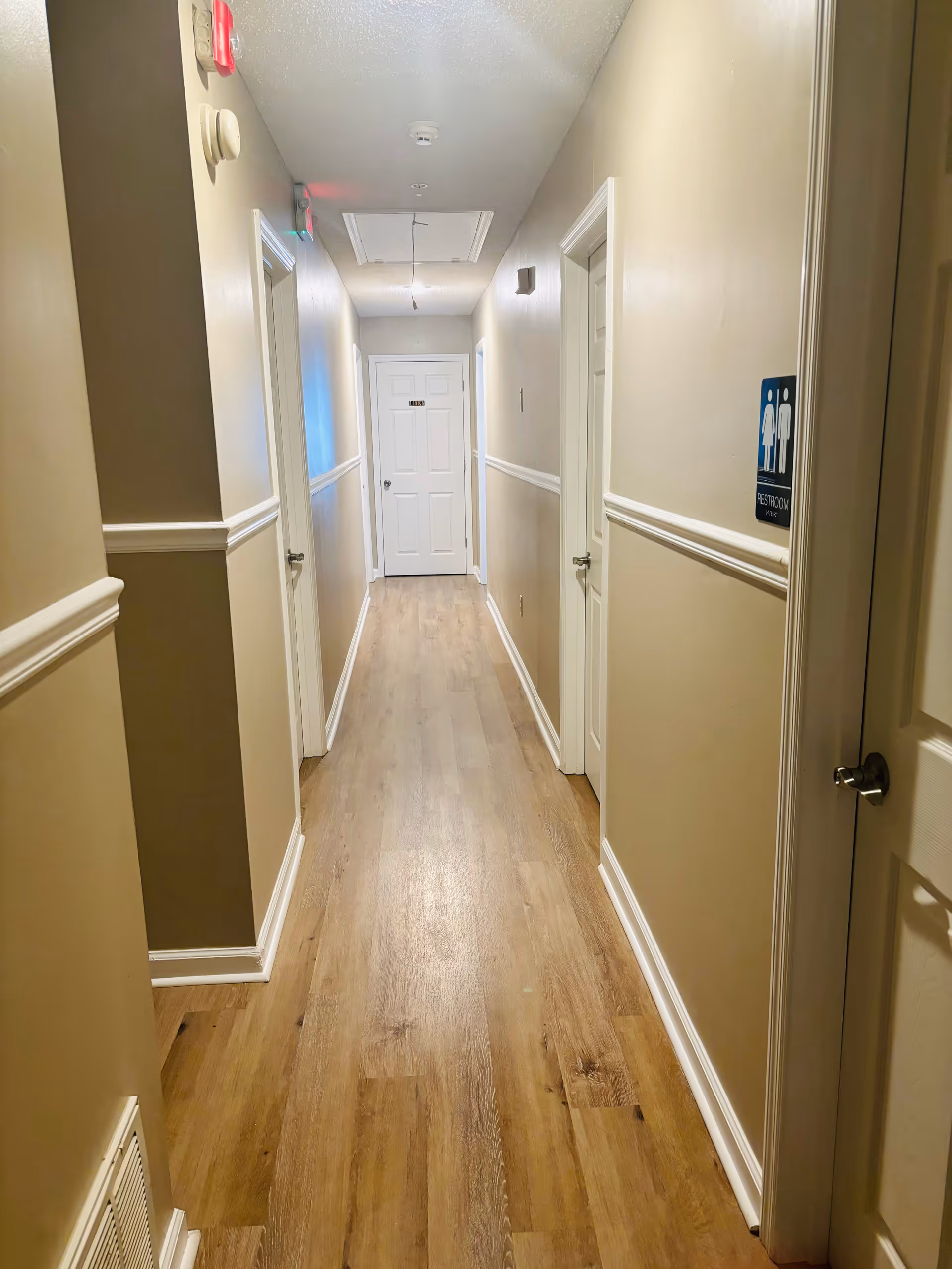 A long, narrow hallway with light beige walls and wood flooring. Several white doors line both sides of the hallway, one of which has a restroom sign indicating male and female restrooms. The ceiling has a smoke detector and an access panel with a hanging wire. The hallway is well-lit with overhead lighting.