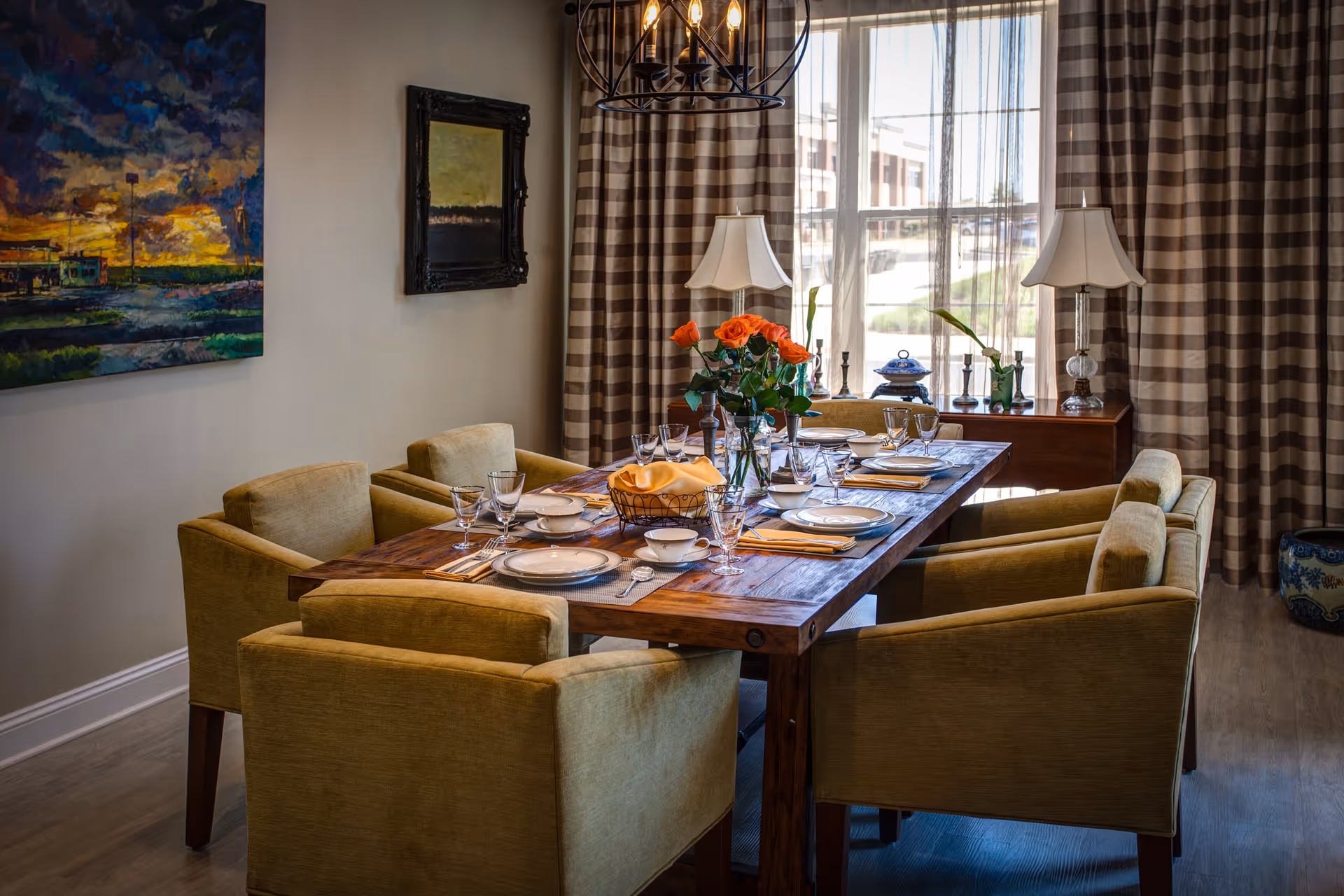 A dining room with a wooden table set for six people. The table is adorned with plates, bowls, glasses, cutlery, napkins, and a centerpiece of orange roses in a vase. Surrounding the table are six upholstered chairs in a muted green color. The room features large windows with sheer and striped curtains, two table lamps on a sideboard, and two paintings on the walls.