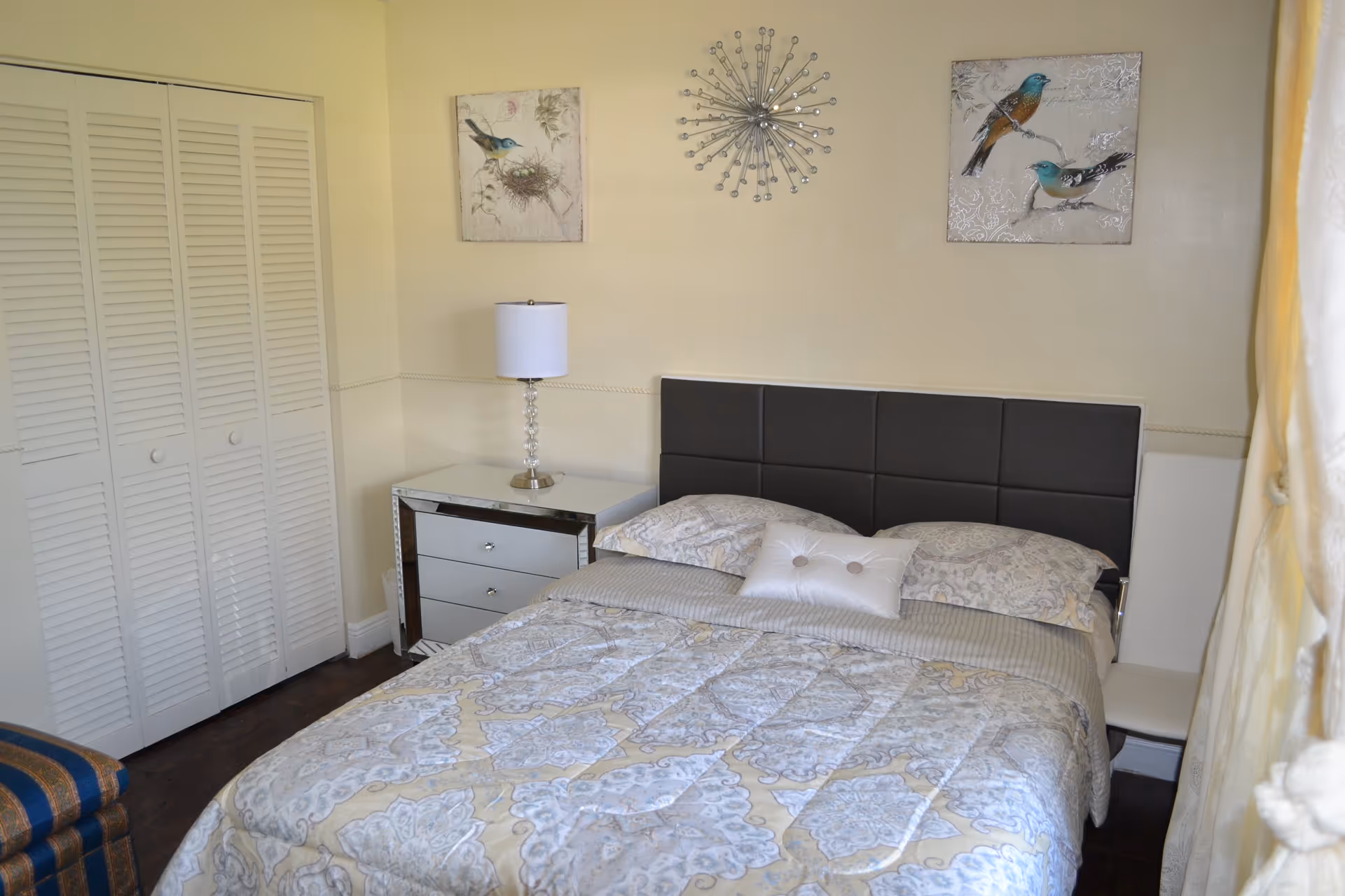 A neatly made bed with patterned bedding, a dark upholstered headboard, bedside table and lamp, closet doors and bird wall art in a small bedroom.