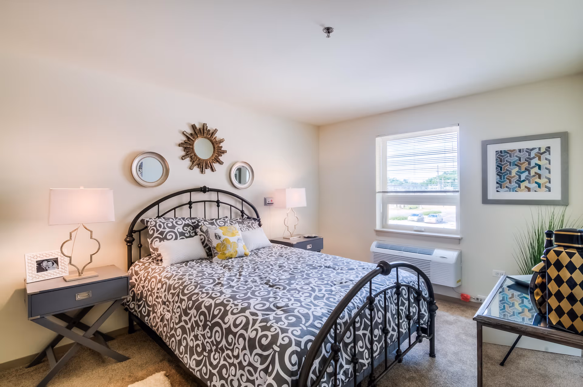 A neatly made bed with a black metal frame and patterned gray and white bedding in a bright bedroom. There are two matching nightstands with lamps on either side of the bed, decorative mirrors on the wall above the headboard, a window with blinds letting in natural light, and a framed geometric artwork on the adjacent wall. A small table with decorative items is positioned near the window.
