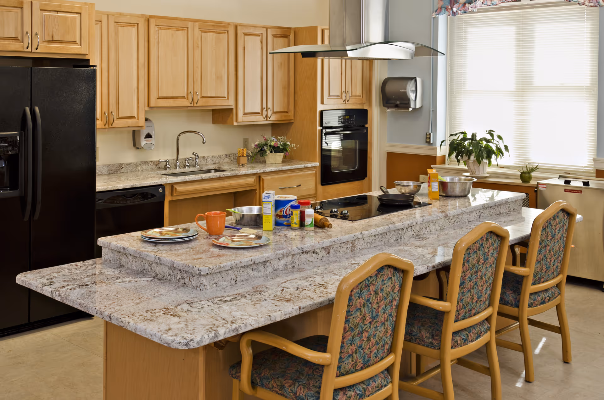 A kitchen area with light wood cabinets, a black refrigerator, a built-in oven, and a granite countertop island with a cooktop. Three wooden chairs with floral upholstery are placed at the island. Various kitchen items including plates, a mug, bowls, and cooking ingredients are on the countertop. A window with blinds and a potted plant is visible in the background.