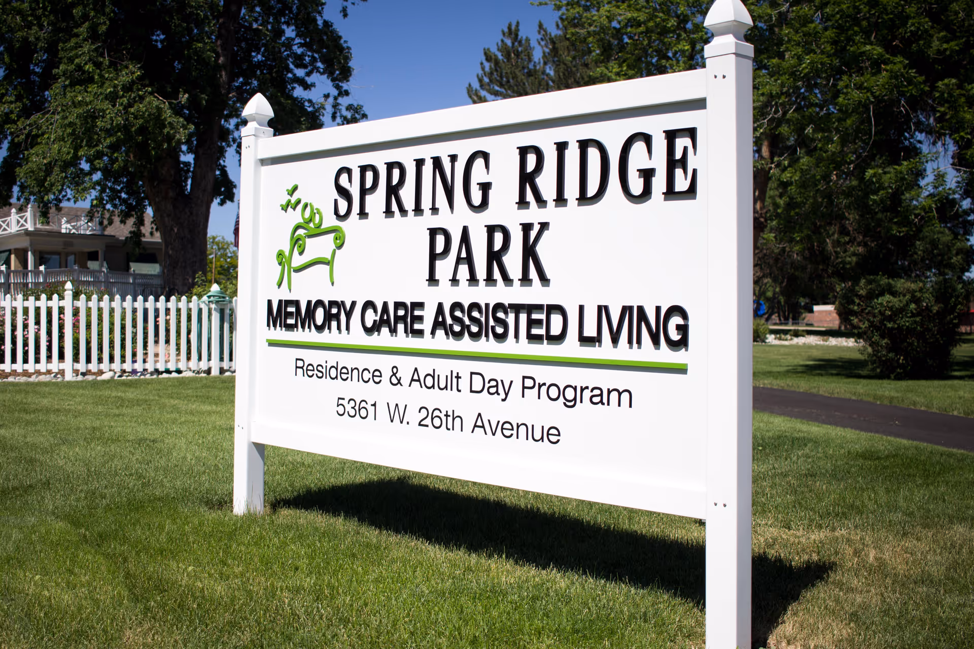 White outdoor sign for Spring Ridge Park Memory Care Assisted Living with address 5361 W. 26th Avenue, set on a grassy area with trees and a white picket fence in the background.