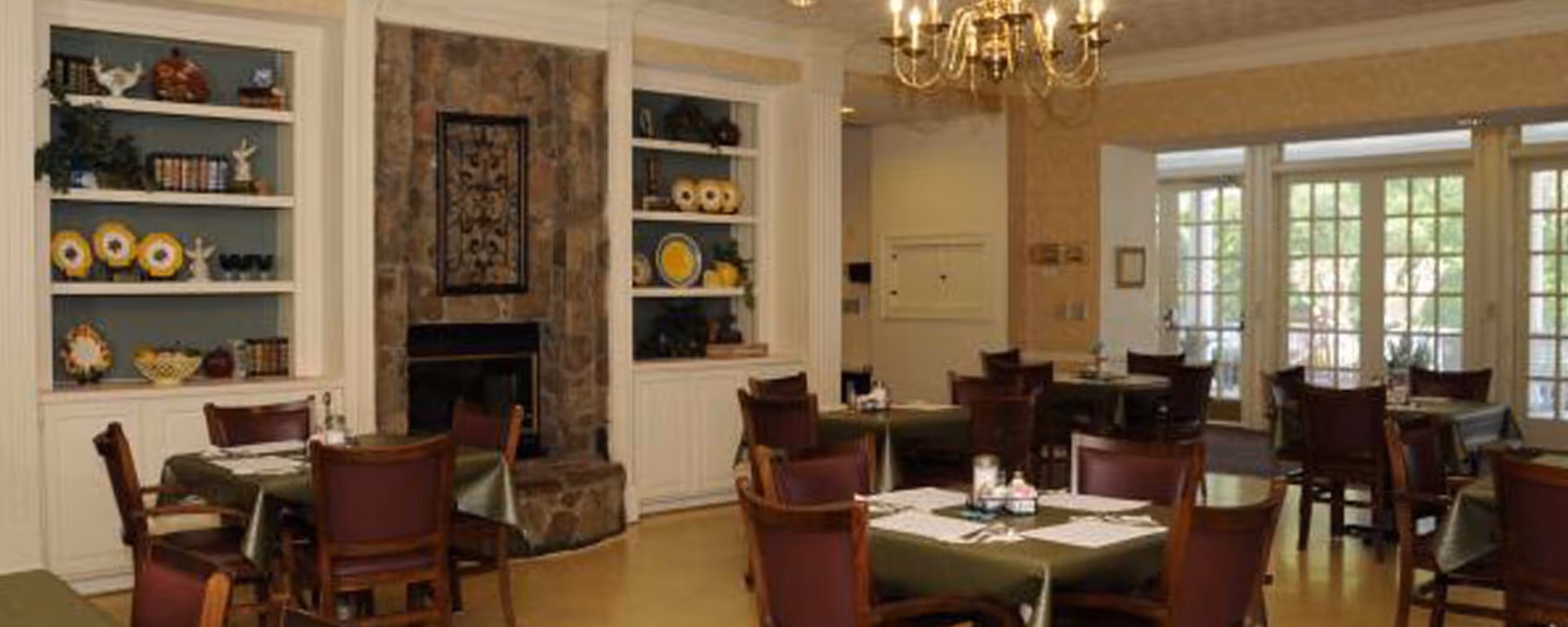Dining room with several tables covered with green tablecloths and set with placemats, utensils, and condiments. The room features wooden chairs, built-in white shelves with decorative plates and books, a stone fireplace with a decorative metal panel above it, and large windows with glass doors letting in natural light. A chandelier hangs from the ceiling.