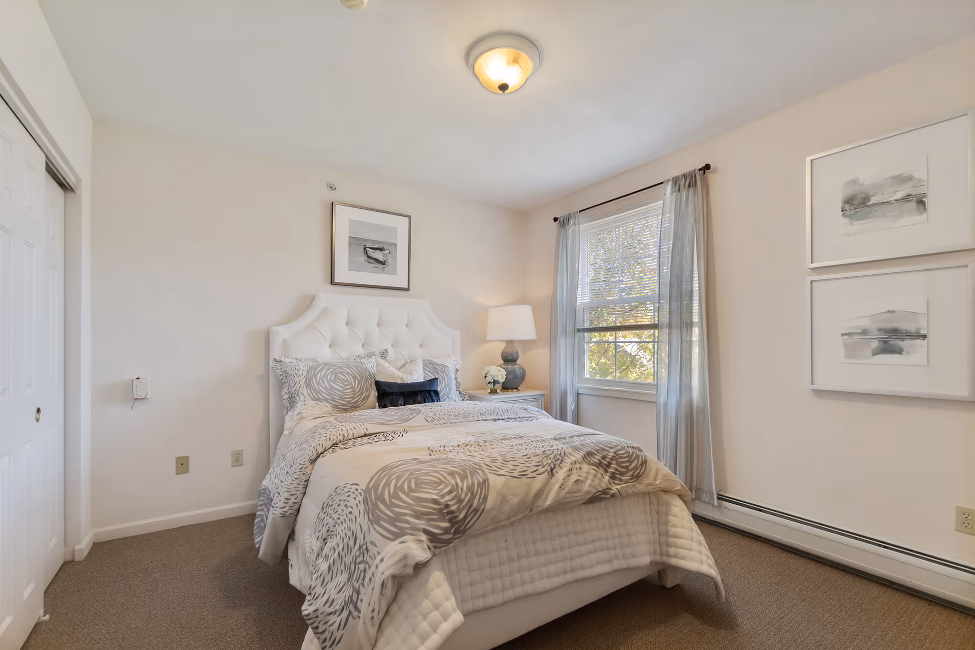 A cozy bedroom with a white tufted headboard bed dressed in a beige and gray patterned comforter and pillows. There is a nightstand with a lamp and a small flower arrangement next to a window with sheer gray curtains. The walls are light-colored with three framed abstract artworks hanging on them. The room has beige carpeting and a ceiling light fixture.