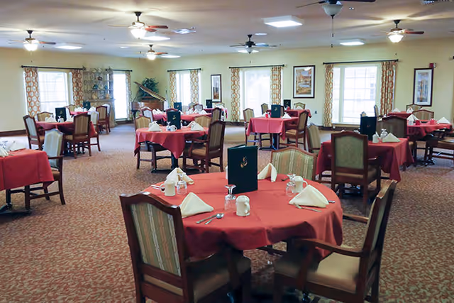 A spacious dining room with multiple round and square tables covered with red tablecloths. Each table is set with white napkins, cups, and silverware. The room has large windows with patterned curtains, ceiling fans, and framed artwork on the walls.