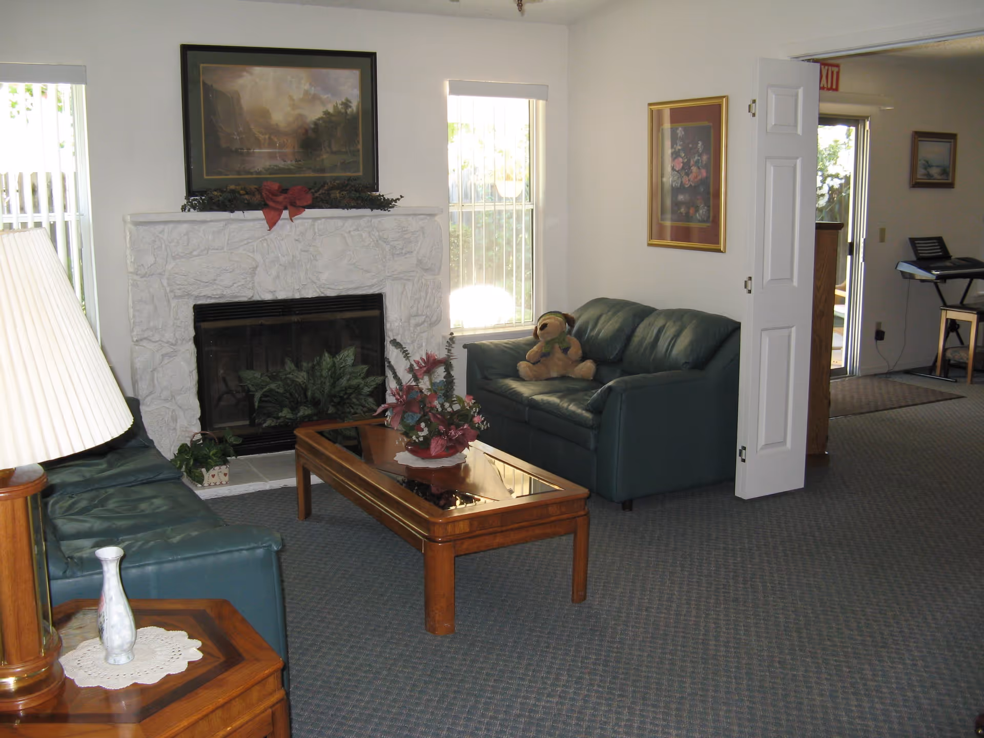 A cozy living room with two green leather sofas, a wooden coffee table with a floral arrangement, a white stone fireplace decorated with greenery and a red bow, and framed paintings on the walls. There is a stuffed dog toy on one of the sofas, a lamp on a wooden side table, and windows letting in natural light.