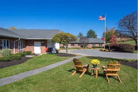 Outdoor view of a senior living facility with a well-maintained lawn, two wooden chairs and a small table with a yellow flower pot in the foreground, a paved walkway, brick buildings in the background, an American flag on a flagpole, and clear blue sky.