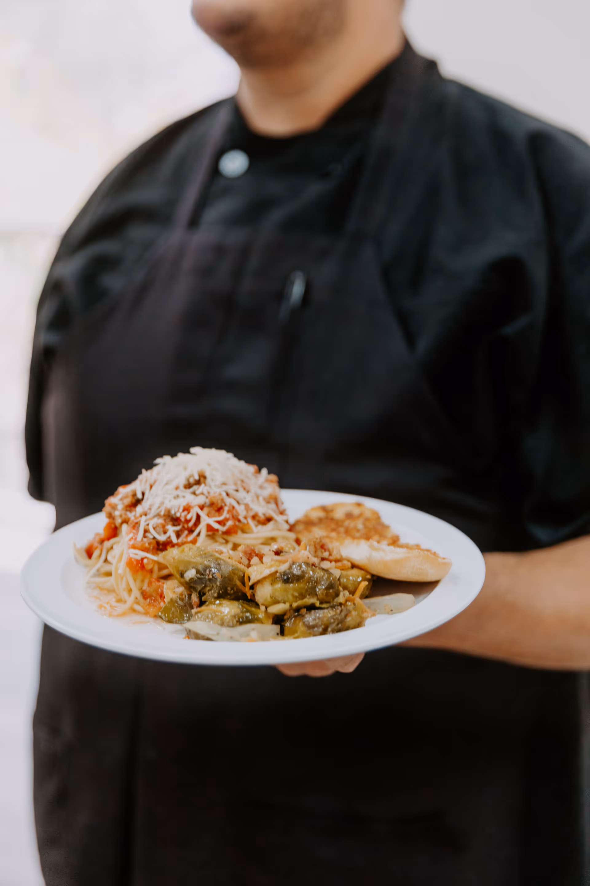 A person wearing a black apron and black shirt holding a white plate with a serving of spaghetti topped with tomato sauce and shredded cheese, cooked vegetables, and a piece of bread.