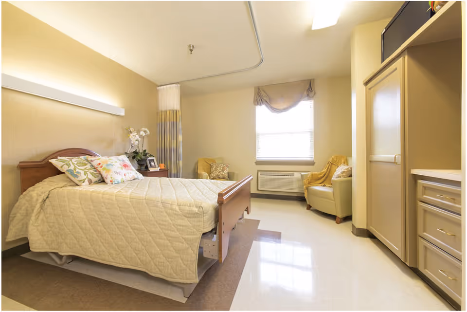 A well-lit nursing facility bedroom with a neatly made bed covered in a beige quilt. The room features a wooden headboard, two floral pillows, a nightstand with a plant and picture frame, a window with a beige curtain, an air conditioning unit below the window, and two comfortable chairs with cushions. There is also a beige cabinet and drawers on the right side of the room.