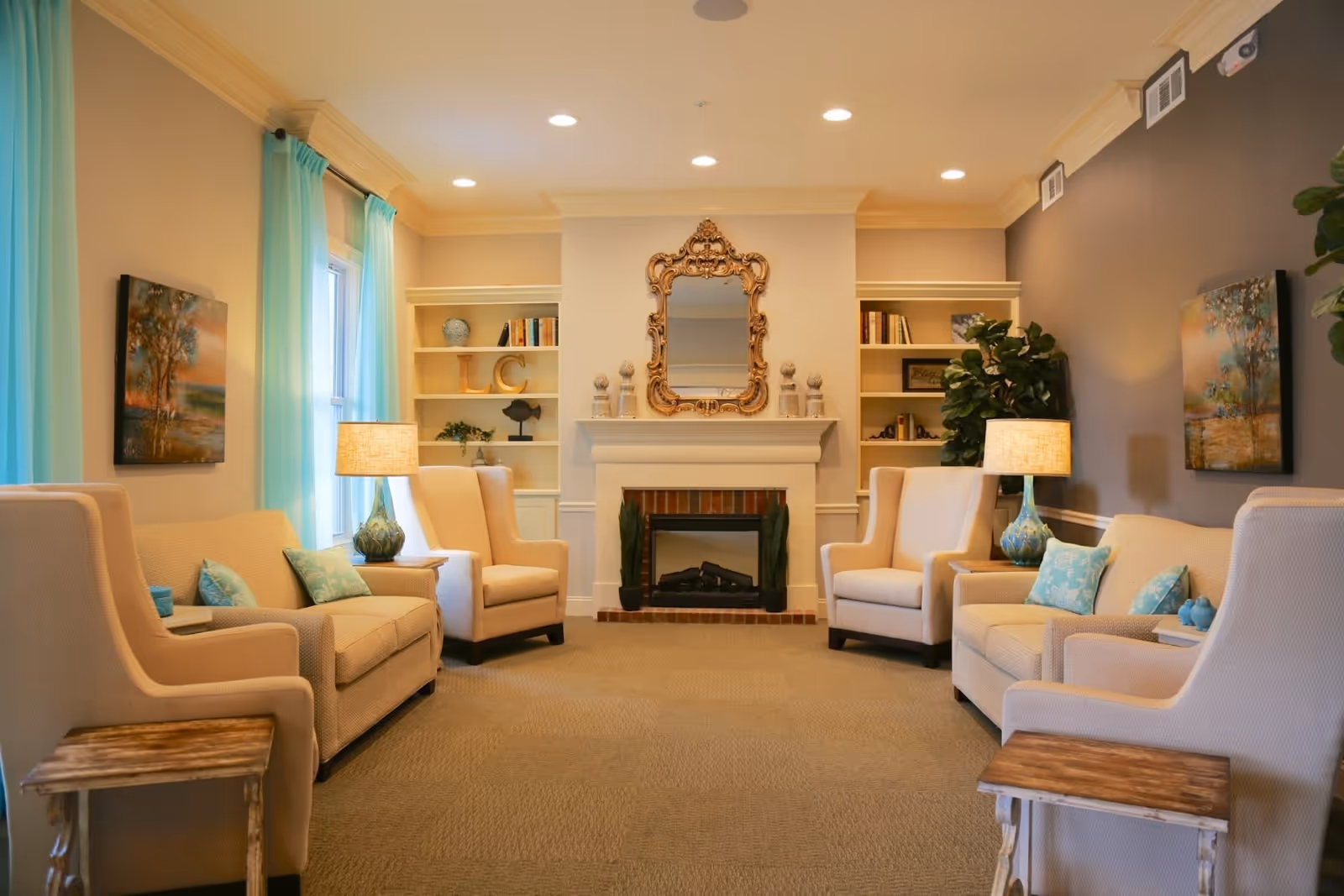 A cozy senior living facility common area with beige armchairs and sofas arranged around a fireplace. The room features light blue curtains, two table lamps with blue bases, built-in shelves with decorative items and books, a large ornate mirror above the fireplace, and framed artwork on the walls.