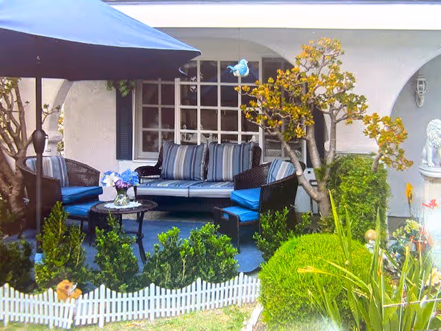 Outdoor seating area with a striped cushioned sofa and chairs under a large umbrella, surrounded by green bushes, small trees, and a white picket fence in front of a building with arched windows.