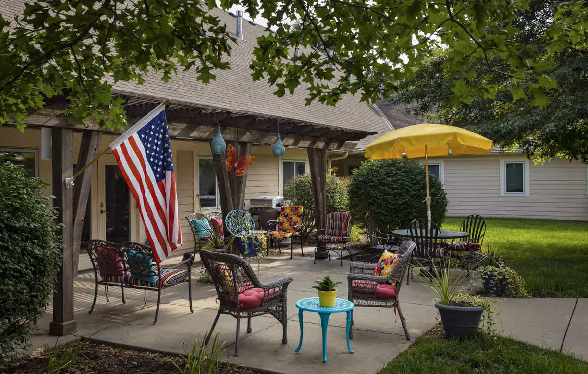 Outdoor patio area at Homestead Assisted Living of Shawnee with various chairs and tables, including a small turquoise table with a potted plant, a yellow umbrella providing shade, and an American flag hanging from a wooden pergola. The patio is surrounded by greenery and bushes, with a beige building in the background.