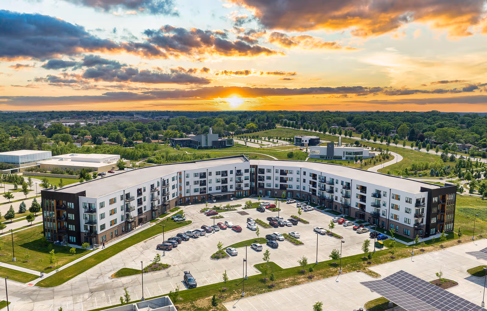 Aerial view of a modern, U-shaped apartment building with multiple floors and balconies, surrounded by parking lots and green spaces, under a partly cloudy sky at sunset.