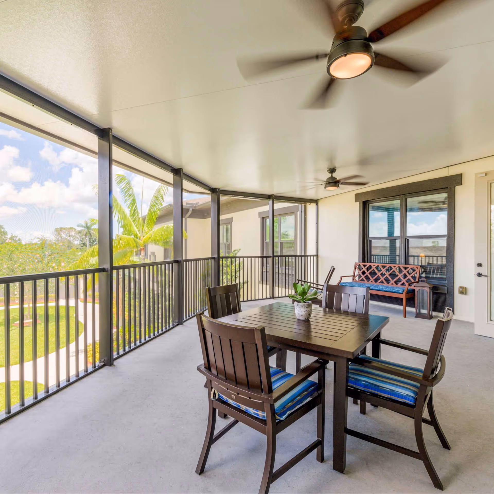 Covered outdoor patio area with a dark wooden table and four chairs with blue striped cushions. There is a small potted plant on the table. The patio has black metal railings and ceiling fans. In the background, there is a wooden bench with a blue cushion and a window with black trim. Palm trees and a clear sky are visible outside.