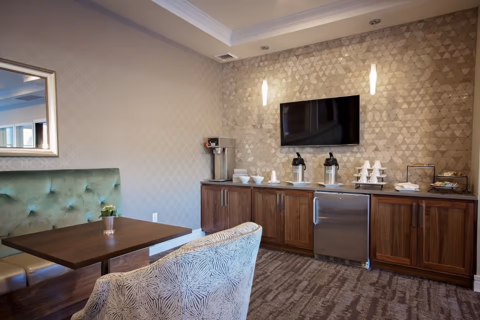 A cozy lounge area with a green tufted bench and a patterned armchair around a wooden table with a small potted plant. Opposite the seating is a wooden cabinet with a coffee station, including coffee dispensers, cups, bowls, and a small refrigerator. A flat-screen TV is mounted on a textured wall above the cabinet, illuminated by two vertical wall lights.