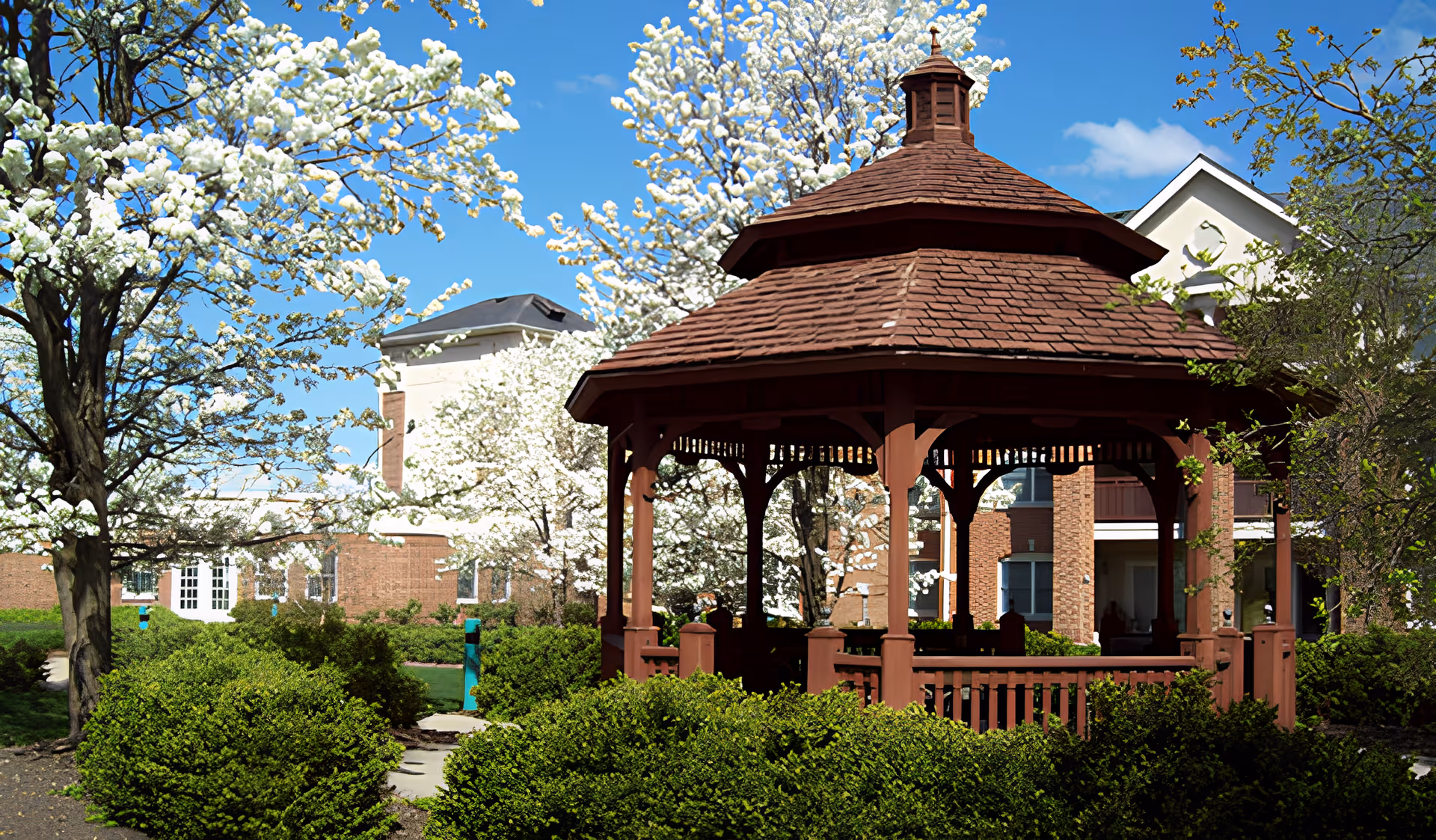 A wooden gazebo surrounded by blooming trees and landscaped shrubs in a senior living facility courtyard.