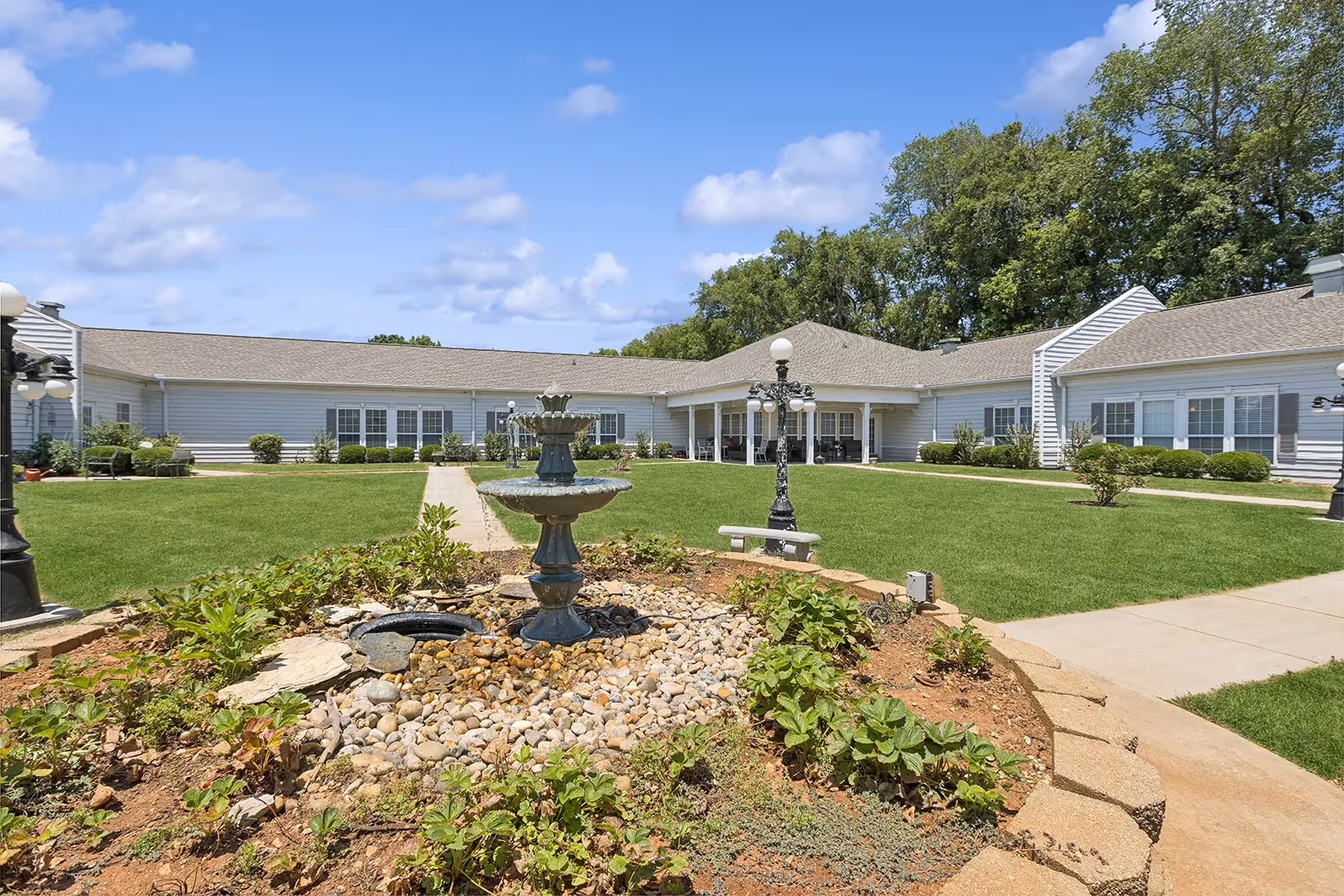 Outdoor courtyard area of The Neighborhood at Sheffield featuring a central multi-tiered water fountain surrounded by landscaping with small plants and rocks. The courtyard is bordered by a single-story building with white siding and multiple windows. There are paved walkways, green grass, and vintage-style black lamp posts under a partly cloudy blue sky.