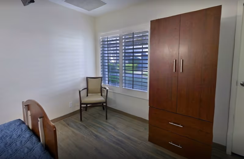 Small bedroom with a single bed, wooden wardrobe, and a chair by a window with shutters.