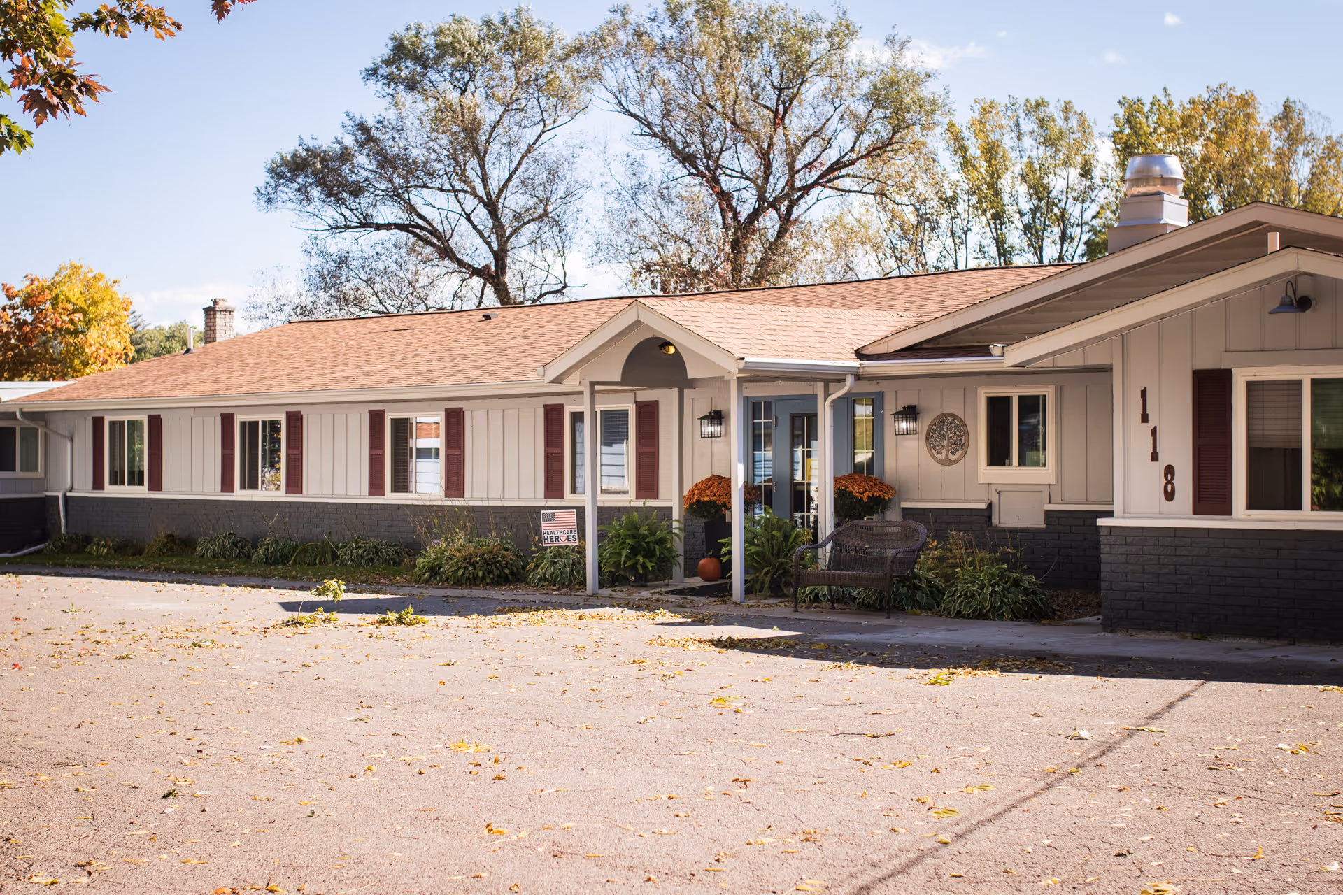 Exterior view of a single-story senior living facility building with beige siding, brown shutters, and a brown roof. The entrance is covered by a small porch with two outdoor wall lanterns and decorated with potted plants and flowers. The building number 118 is visible on the right side near a window. Trees with autumn foliage are in the background under a clear sky.