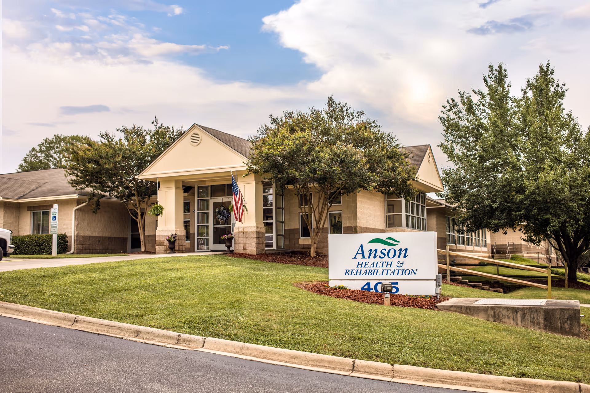 Exterior view of Anson Health & Rehabilitation building with a well-maintained lawn, trees, and an American flag near the entrance. The facility sign is visible on the grass in front of the building.