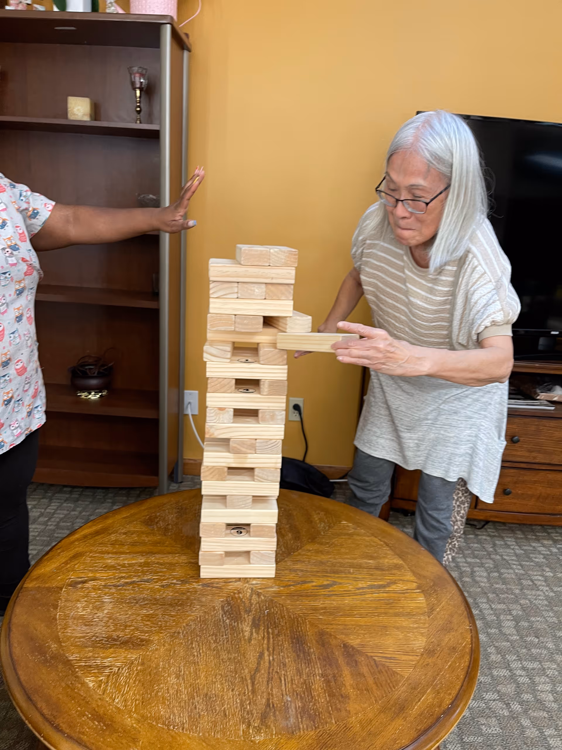 An elderly woman with gray hair and glasses is playing a game of Jenga on a wooden round table. Another person, partially visible, is standing nearby with their arm extended. The setting appears to be a cozy living room with a wooden cabinet, a television, and a yellow wall in the background.