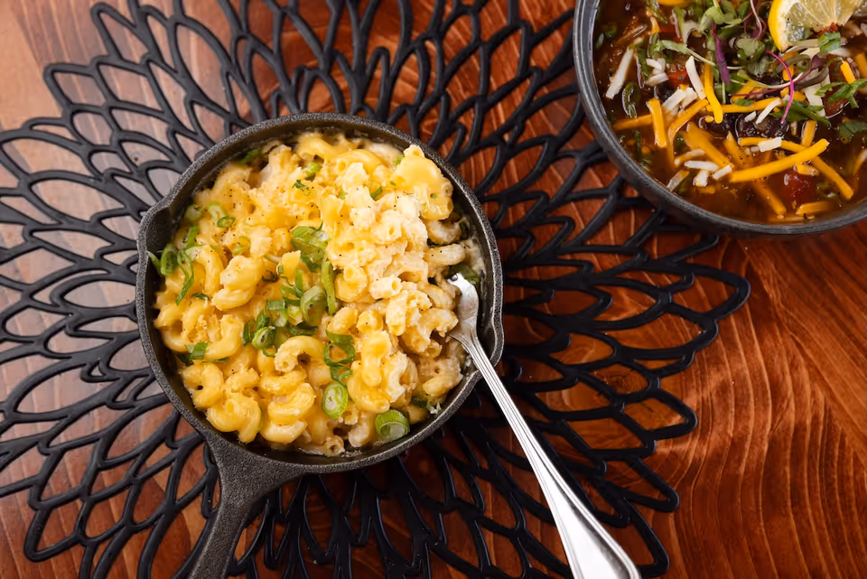 A small cast iron skillet filled with macaroni and cheese garnished with chopped green onions, placed on a decorative black placemat on a wooden table. Next to it is a bowl of soup topped with shredded cheese, chopped onions, microgreens, and a lemon wedge.