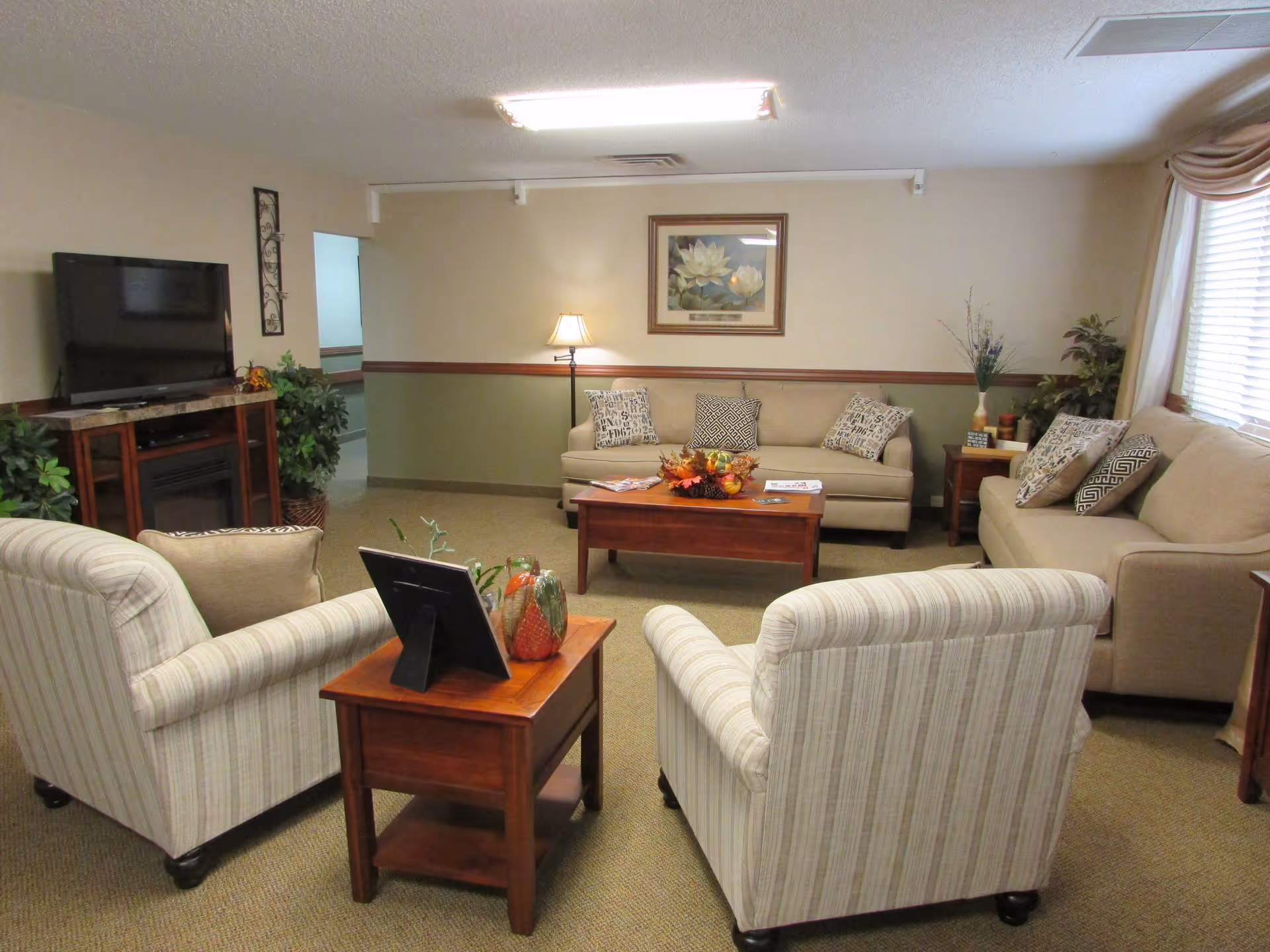 A cozy living room in a senior living facility featuring two beige sofas with patterned cushions, two striped armchairs, a wooden coffee table with a fall-themed centerpiece, a side table with decorative items, a floor lamp, a flat-screen TV on a wooden stand, and framed artwork on the wall. The room has beige walls with a green lower half and carpeted floor.