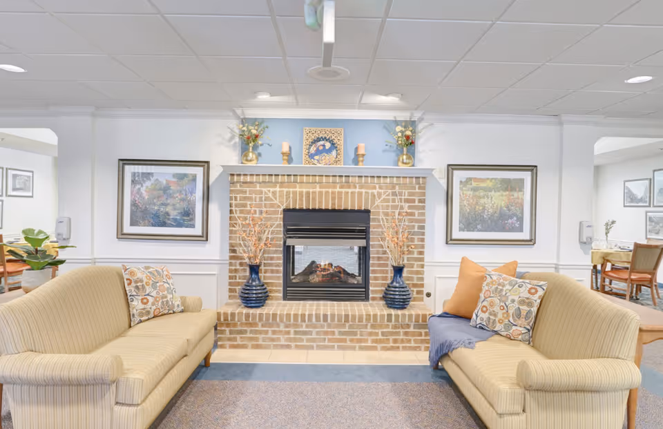 A cozy living room area with two beige striped sofas facing each other in front of a brick fireplace. The fireplace mantel is decorated with two vases holding dried flowers, two candles, and a framed artwork. There are framed paintings on the walls on either side of the fireplace. The room has a light and airy feel with white walls and ceiling tiles.