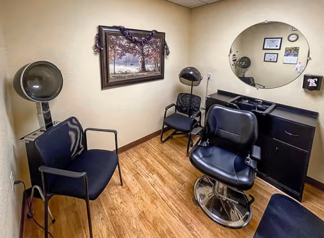Small salon room with a black salon chair, two black waiting chairs, a hair dryer hood, a large round mirror on the wall, a black cabinet with a sink, and a framed picture of a tree on the wall.