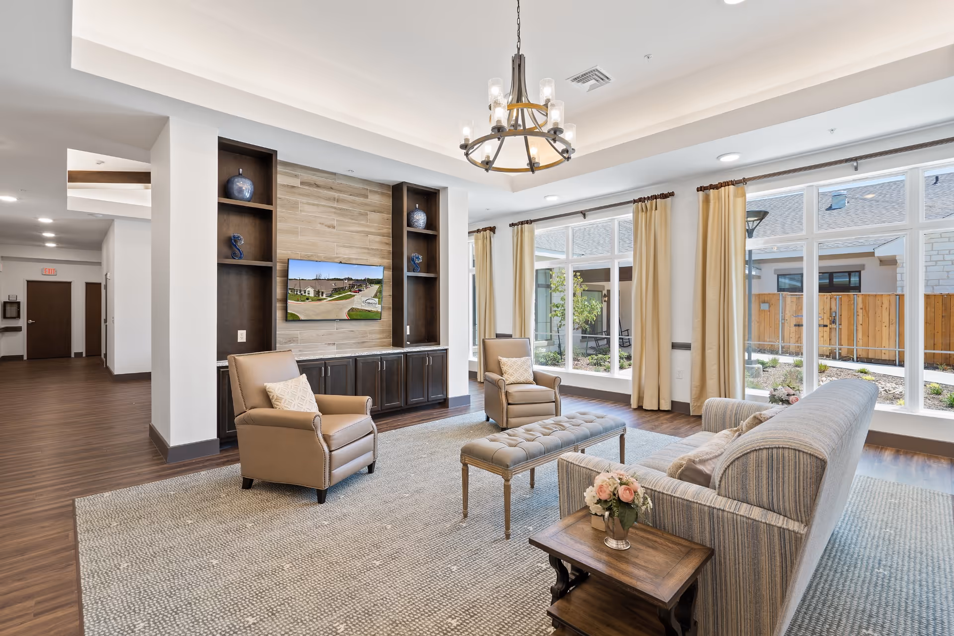 A bright and spacious senior living facility common area with large windows letting in natural light. The room features a striped sofa, two beige armchairs with patterned pillows, a tufted bench, and a wooden side table with a flower arrangement. A flat-screen TV is mounted on a wood-paneled wall with built-in dark wood shelves and cabinets. The flooring is a combination of wood and a large area rug, and a modern chandelier hangs from the ceiling.