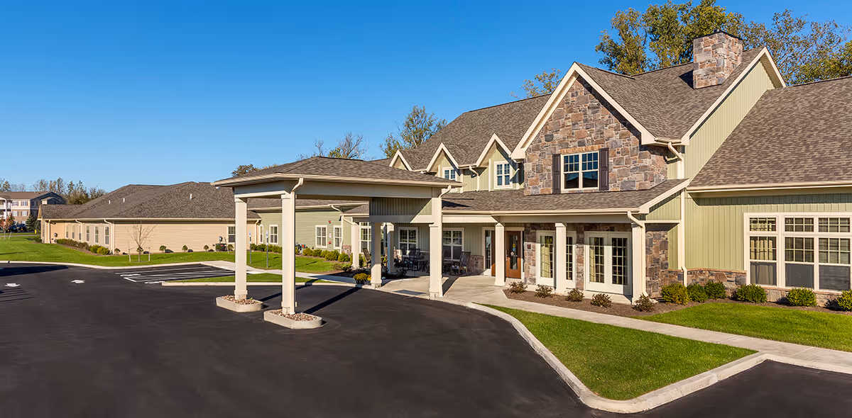 Front entrance of a senior living community building with a covered porte-cochère, stone and siding facade, and adjacent parking lot.