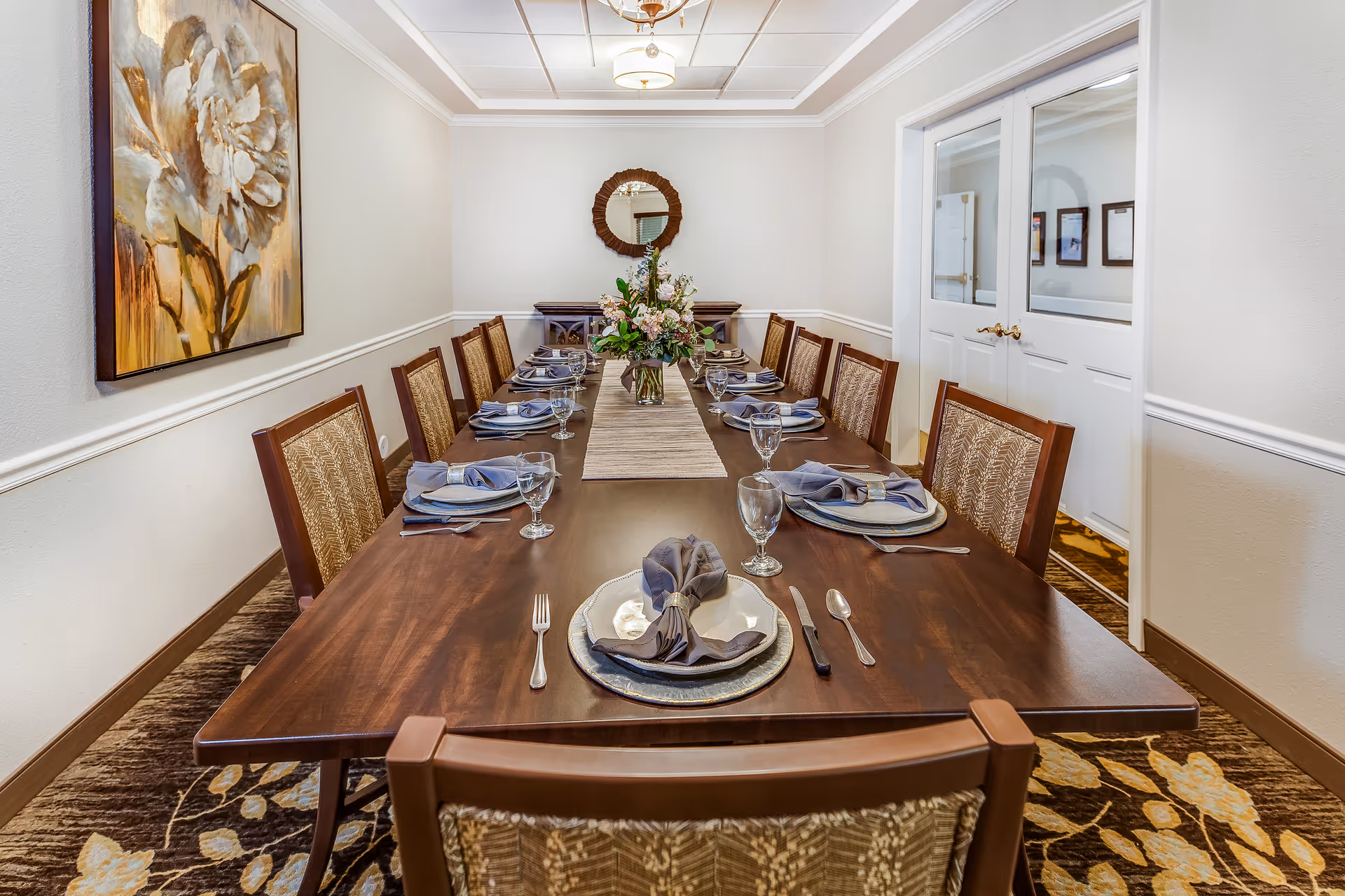 A formal dining room with a long wooden table set for ten people. Each place setting includes a plate, a folded gray napkin with a napkin ring, a fork, knife, spoon, and a water glass. A floral centerpiece is placed in the middle of the table. The room has beige walls, a large floral painting on the left wall, a round mirror on the far wall, and double white doors with glass panels on the right. The floor is carpeted with a floral pattern.