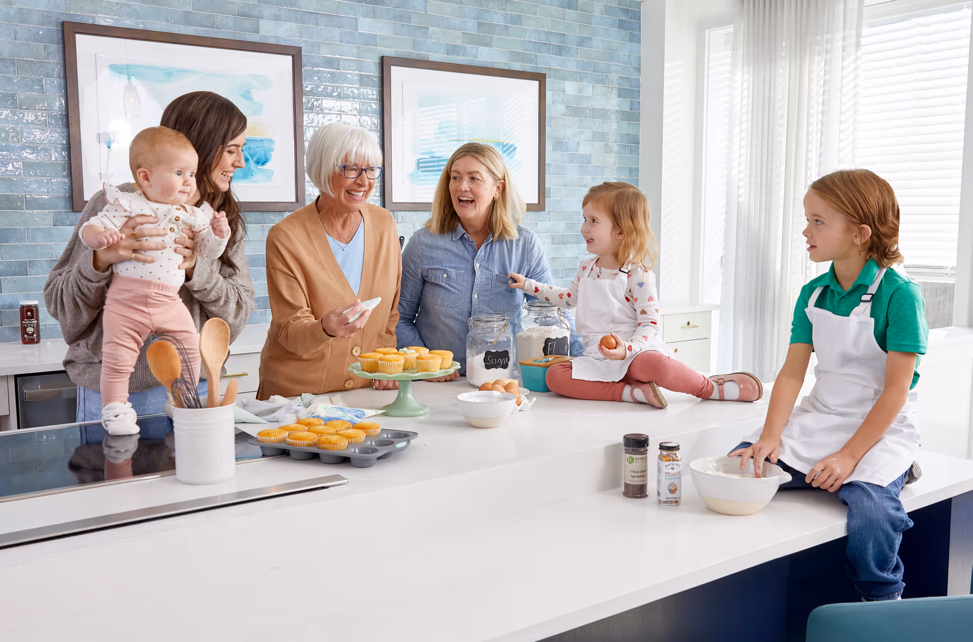 A multi-generational family baking together in a modern kitchen. An older woman and two middle-aged women are smiling and interacting with three young children, one of whom is a baby being held by a woman. The children are wearing aprons and sitting or standing on a white kitchen island with baking ingredients and freshly baked cupcakes.