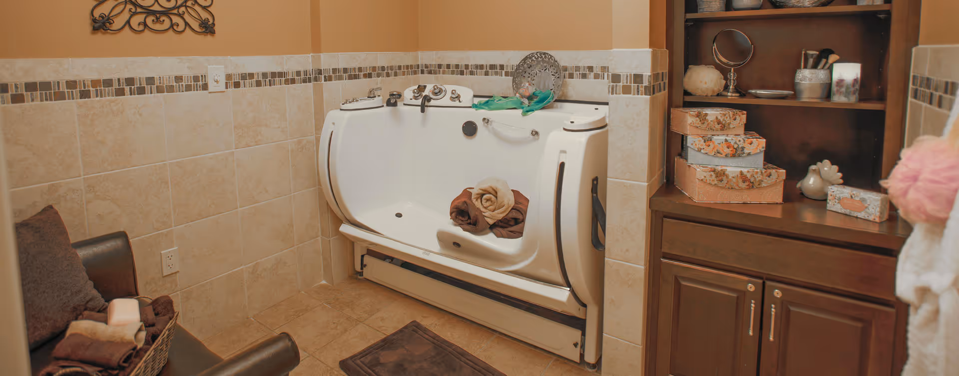 A bathroom with beige tiled walls and floor featuring a white walk-in bathtub with rolled towels inside. To the right, there is a wooden cabinet with decorative boxes and toiletries. On the left, a dark chair with a basket of towels is visible.