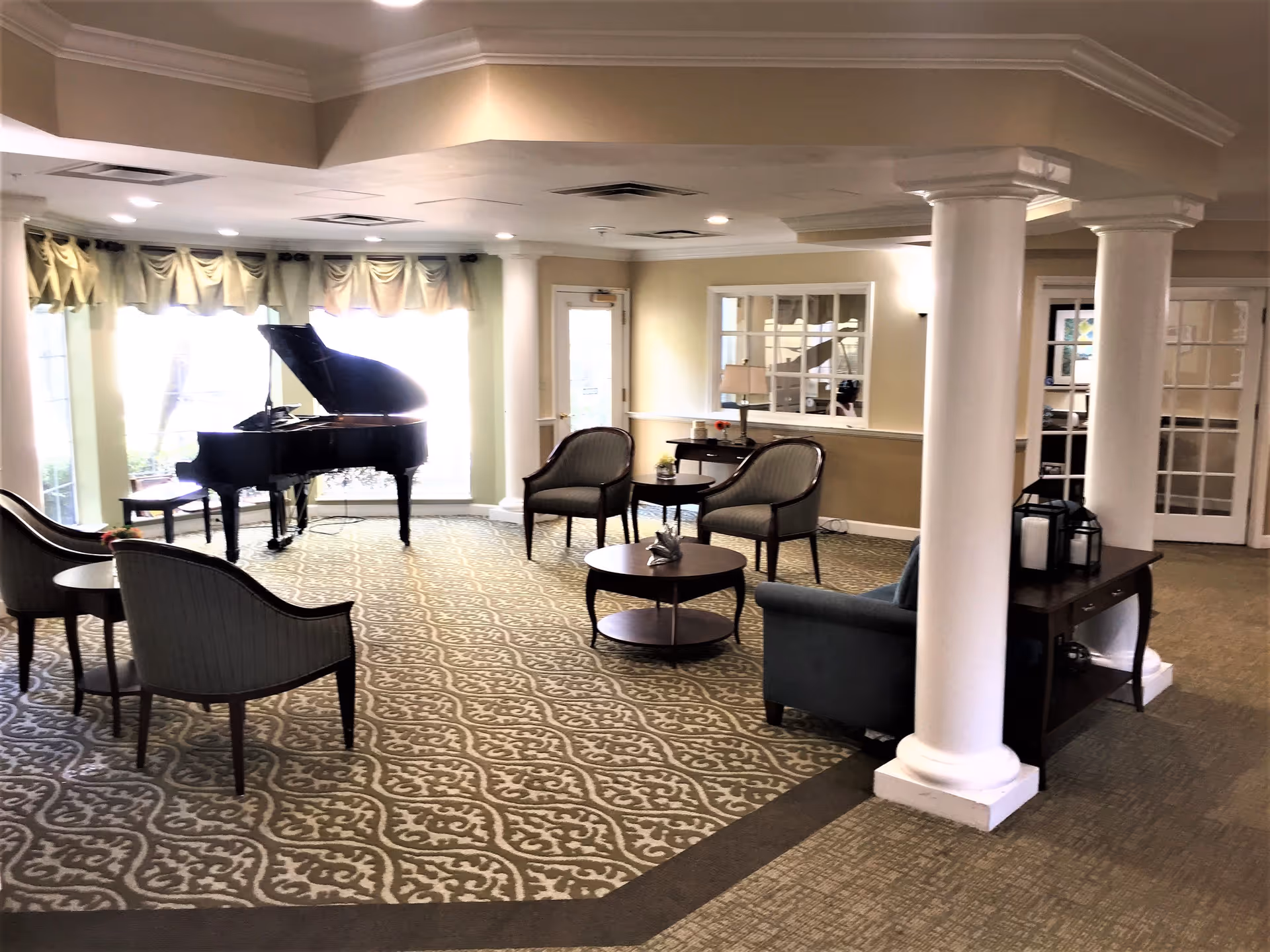 A spacious, well-lit living room area with patterned carpet, several upholstered chairs, a round coffee table, a grand piano near large windows with curtains, white columns, and a console table with decorative lanterns.