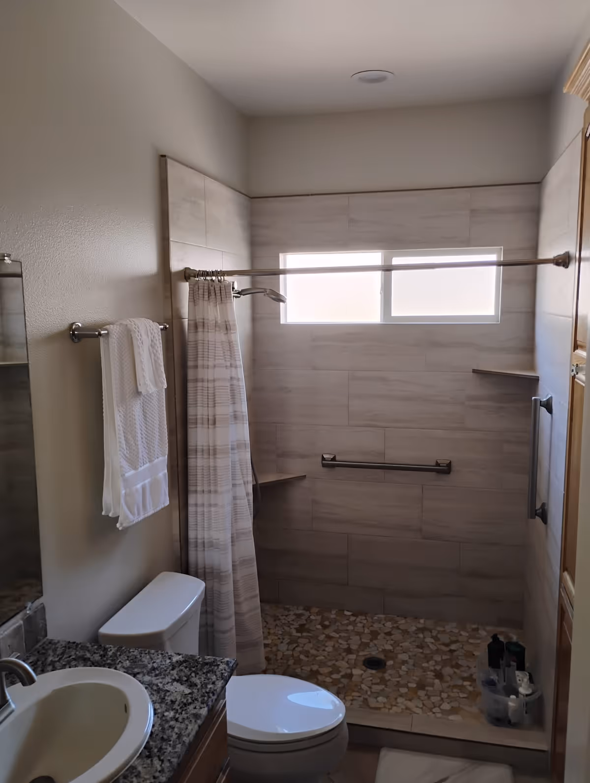 A bathroom with a granite countertop sink, a toilet, and a walk-in shower with a pebble stone floor and tiled walls. The shower has a grab bar, a small corner shelf, a shower curtain, and a frosted window letting in natural light. A towel hangs on a rack next to the toilet.