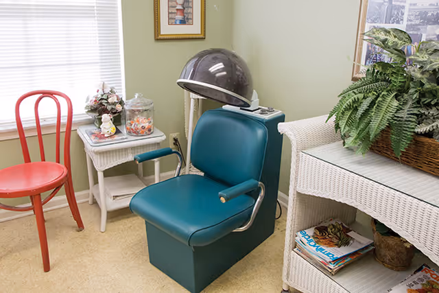 A small room with a vintage hair dryer chair in teal color, a red wooden chair, a white wicker side table with a glass jar of candy and a small decorative item, and a white wicker shelf holding magazines and a large potted fern. A window with blinds and a framed picture on the wall are also visible.