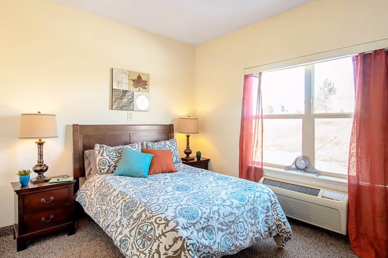 A cozy bedroom with a double bed featuring a patterned blue and white bedspread and three decorative pillows in teal and rust colors. There are two dark wooden nightstands on either side of the bed, each with a lamp and small decorative items. A large window with red curtains lets in natural light, and a clock sits on the windowsill above an air conditioning unit. The walls are painted light beige, and a piece of wall art hangs above the bed.