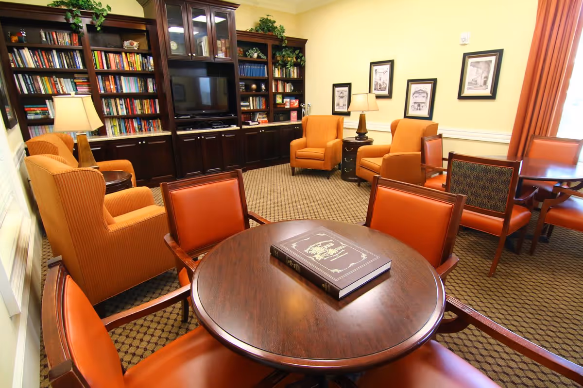 A cozy living room area with orange upholstered armchairs and wooden chairs around a round wooden table with a large book on it. The room features a large dark wood built-in bookshelf filled with books and a flat-screen TV. There are framed pictures on the yellow walls and two table lamps providing warm lighting.