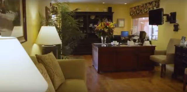 Reception-style living room area with sofa and lamps, a wooden desk topped with flowers, bookshelves and a wall-mounted TV.