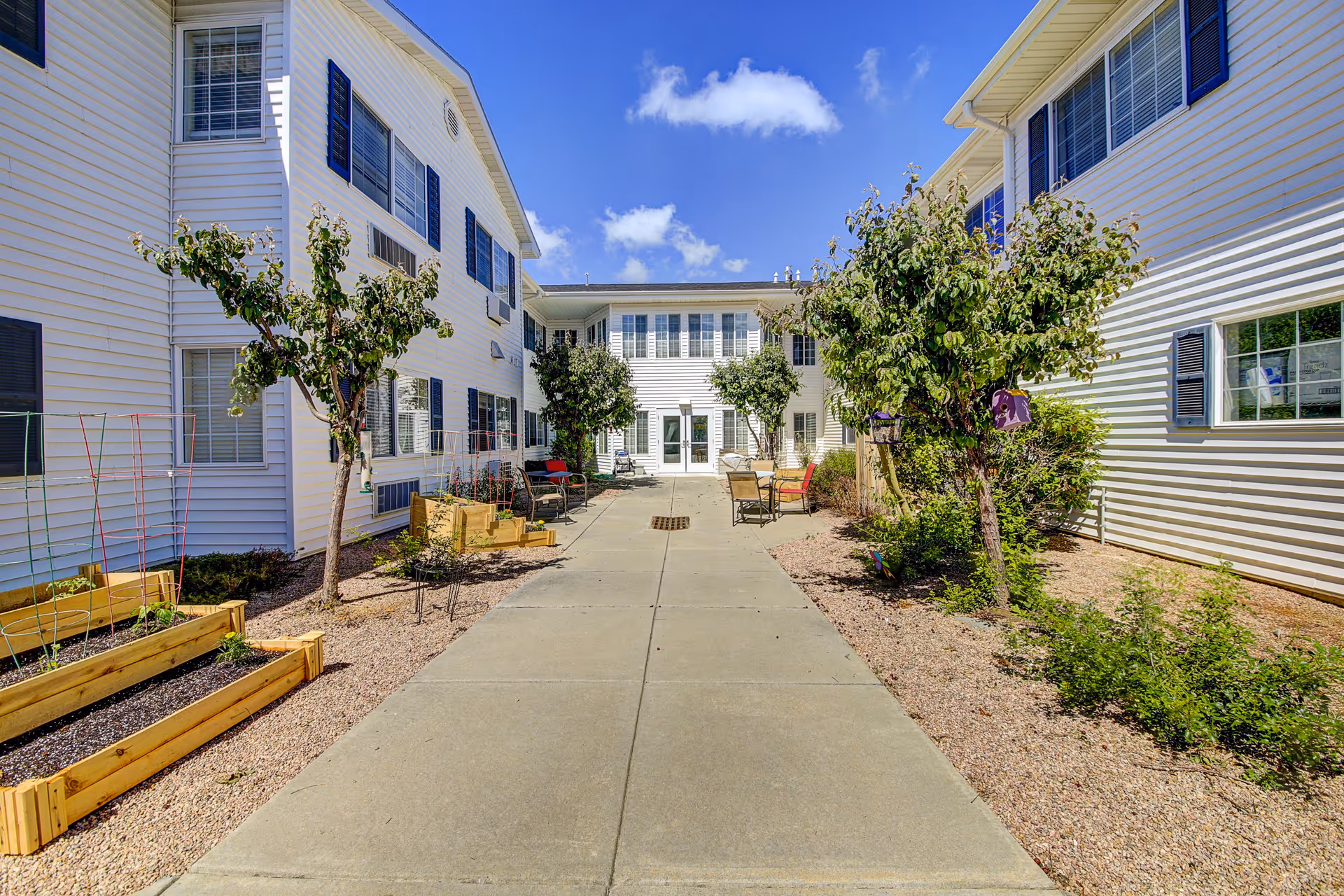 Outdoor courtyard area of Waneka Park Assisted Living with a concrete pathway flanked by small trees, raised garden beds, and seating areas. The courtyard is surrounded by white buildings with blue shutters under a bright blue sky with a few clouds.