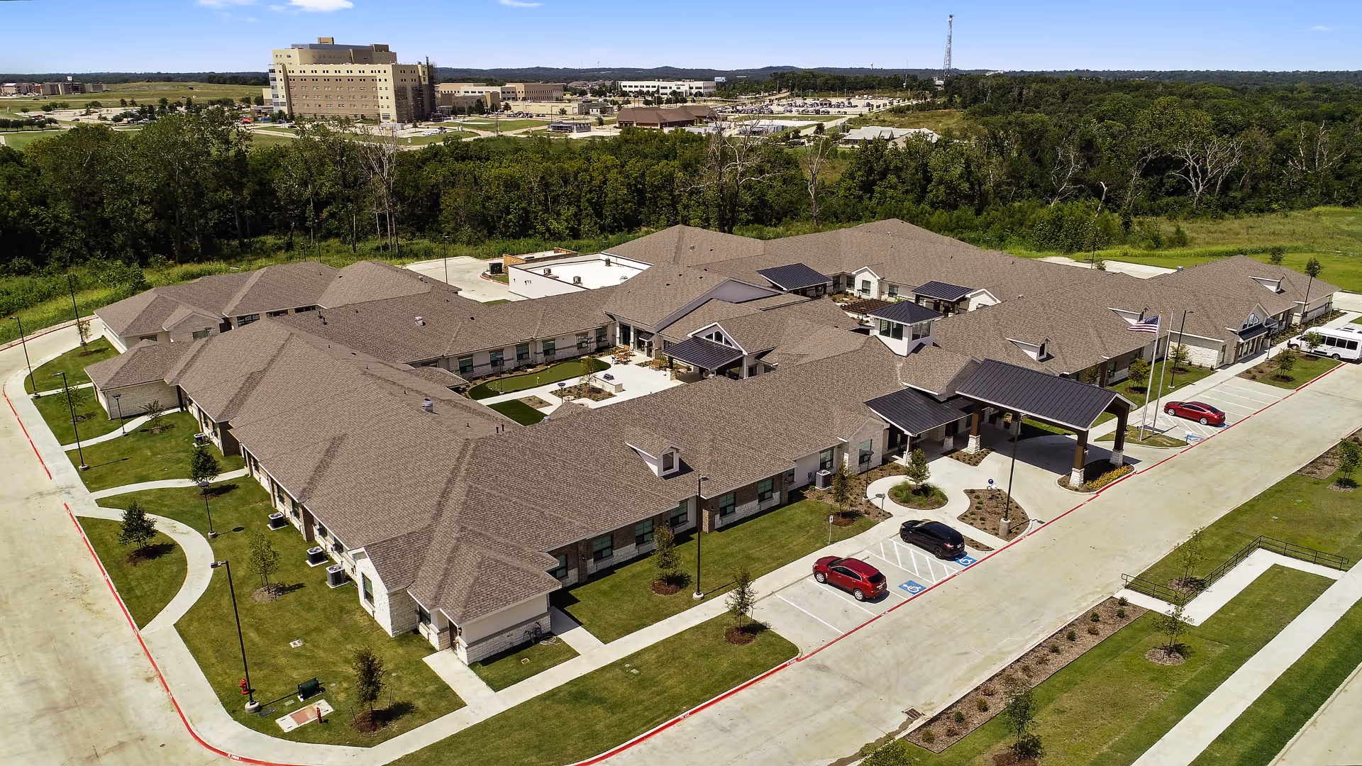 Aerial view of Celebration Senior Living of Denison, showing a large single-story building with a brown roof, surrounded by green lawns, trees, and parking spaces with several cars parked. The building has a covered entrance and is situated near a wooded area with other buildings visible in the distance.