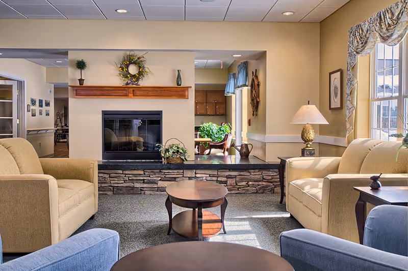 Sunlit communal living room with armchairs, coffee tables, a stone-front fireplace and decorative accents.