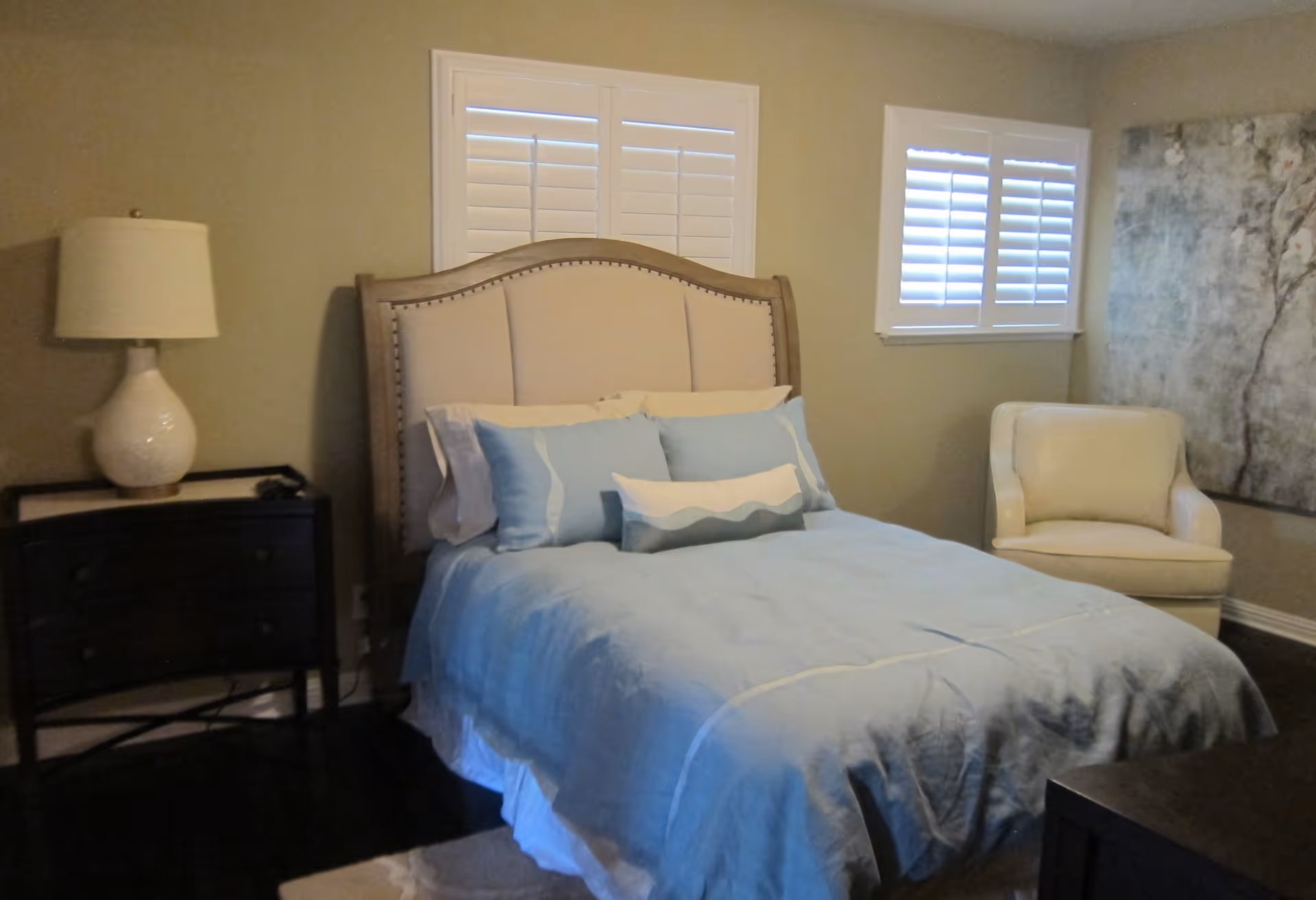 A neatly made bedroom with a bed dressed in light blue bedding, an upholstered headboard, a nightstand with a lamp, and an armchair by a window.
