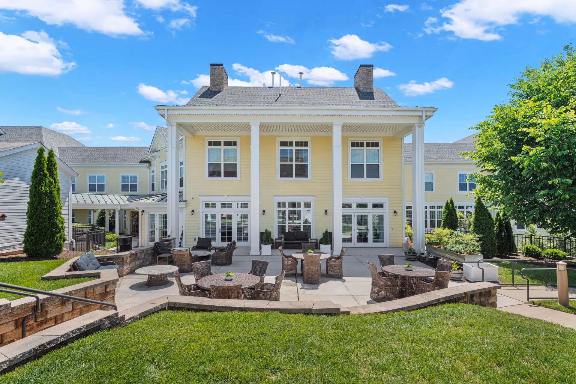 Rear exterior of a yellow two-story senior living building with white columns, an outdoor patio with multiple wicker tables and chairs, lawn and a tree under a blue sky.